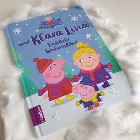Ein Kinderbuch mit dem Titel Peppa Pig und Klara Lina: Fröhliche Weihnachten! liegt auf einer weißen, von Kunstschnee umgebenen Fläche und zeigt Peppa Pig Figuren in festlicher Winterkleidung.