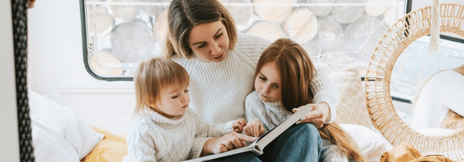 Eine Frau in einem weißen Pullover sitzt mit zwei kleinen Kindern in einem gemütlichen, hellen Raum mit einem großen Fenster und einem Stapel Brennholz, der von draußen zu sehen ist, und liest gemeinsam ein Buch.