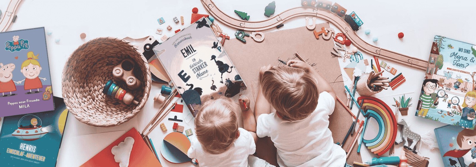 Zwei kleine Kinder sitzen auf dem Boden, umgeben von buntem Spielzeug, Holzeisenbahnschienen, Stiften und mehreren Bilderbüchern, lesen gemeinsam und genießen eine spielerische, kreative Umgebung.