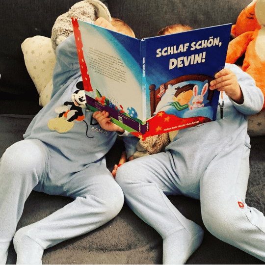 Zwei kleine Kinder in blauen Schlafanzügen sitzen auf einer Couch und lesen ein Kinderbuch mit dem Titel "Schlaf schön, Devin!" Neben ihnen liegt ein Plüschtier, und ihre Gesichter sind größtenteils durch das Buch verdeckt.