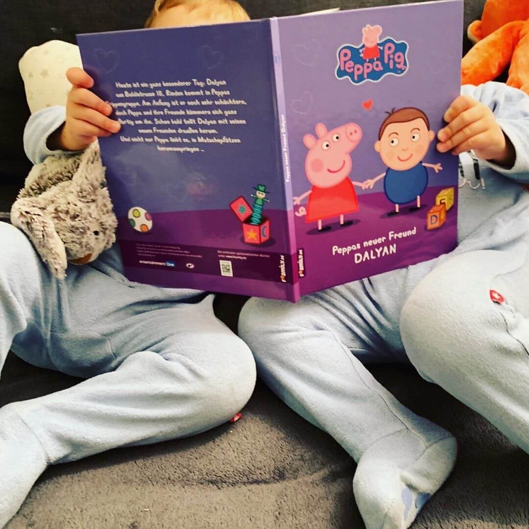 Zwei Kinder in blauen Schlafanzügen sitzen auf einer Couch und lesen ein Peppa Pig-Buch mit dem Titel "Peppas neuer Freund DALYAN". Ein Kind hält ein Plüschhäschen in der Hand, und ihre Gesichter sind hinter dem Buch verborgen.