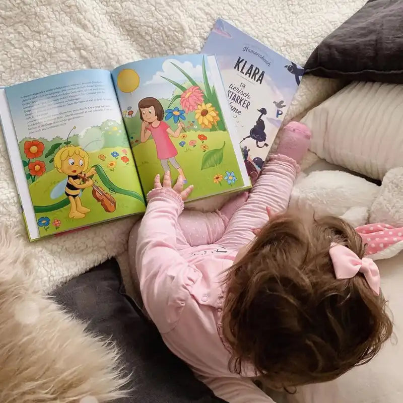 Ein kleines Kind mit einer rosafarbenen Schleife und einem rosafarbenen Schlafanzug sitzt auf einem gemütlichen Bett und betrachtet das personalisierte Framily-Buch "Biene Maja und du", das Biene Maja und ein Mädchen im Garten zeigt. Ein anderes Buch liegt in der Nähe.