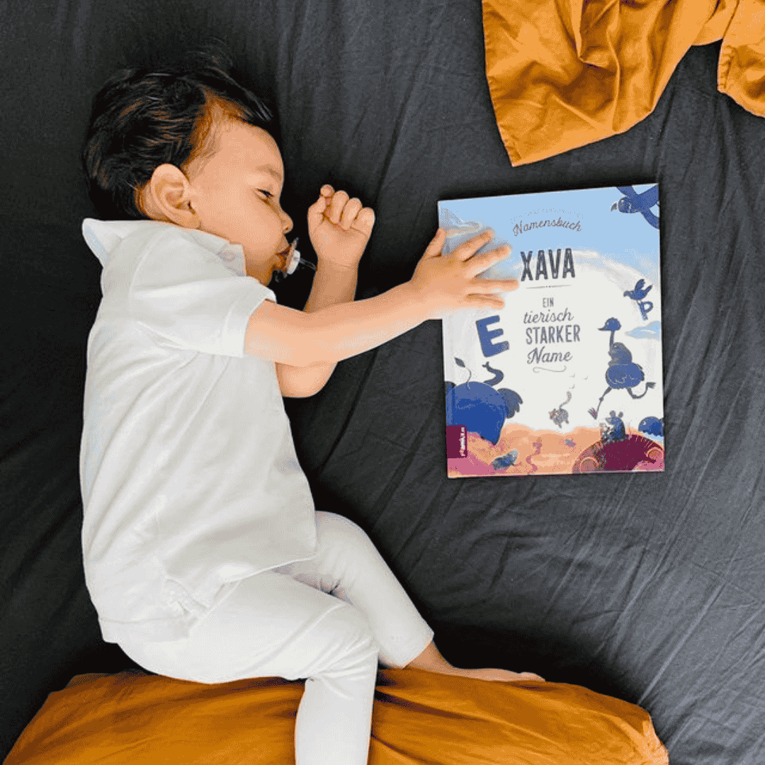Ein weiß gekleidetes Kleinkind schläft in einem dunklen Bett, hält einen Schnuller in der Hand und stützt sich mit einer Hand auf das bunte Kinderbuch Xava Ein wirklich starker Name. Eine orangefarbene Decke liegt in der Nähe und macht die Szene noch gemütlicher.
