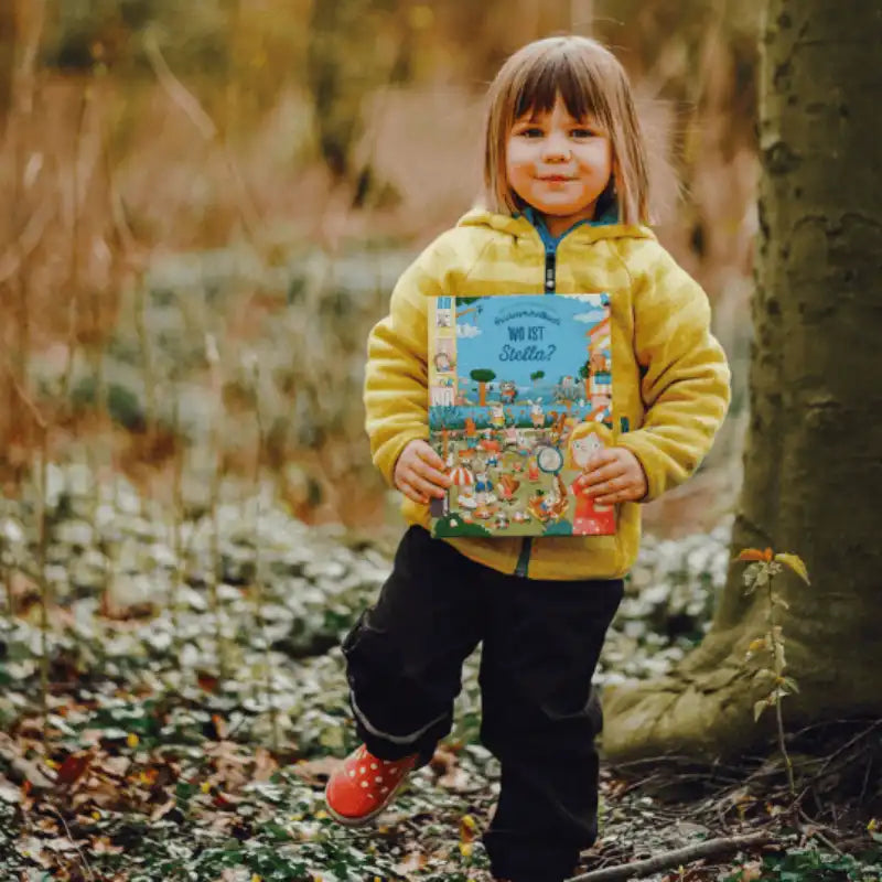 Kind neben einem Baum mit einem Wimmelbuch n der Hand.