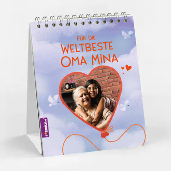Der Framily Tischkalender weltbeste Oma zeigt einen spiralgebundenen Fotokalender mit einem herzförmigen Rahmen mit einer lächelnden älteren Frau und einem jungen Mädchen. "Für die weltbeste Oma Mina" steht auf einem lila Hintergrund mit weißen Schmetterlingen.
