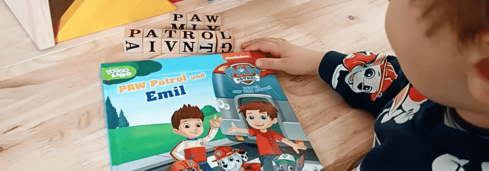Ein Kind im PAW-Patrol-Shirt greift nach einem PAW-Patrol-Buch mit dem Titel "PAW Patrol und Emil", das auf einem Holztisch liegt. In der Nähe befinden sich hölzerne Buchstabenstempel mit der Aufschrift PAW PATROL.