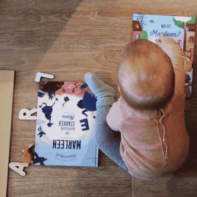Ein Baby sitzt auf einem Holzboden und greift nach einem farbenfrohen Bilderbuch, während ein anderes Buch und Holzbuchstaben, die ART buchstabieren, in der Nähe liegen.