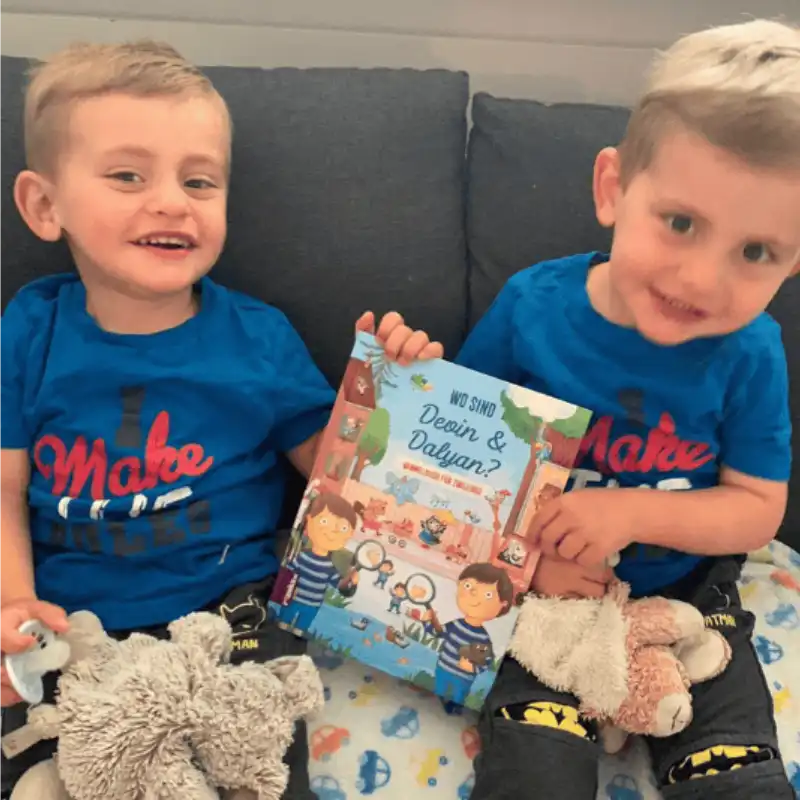Zwei kleine Jungen mit kurzen Haaren und blauen Hemden sitzen lächelnd auf einem Sofa, jeder hält ein Stofftier und zeigt stolz sein Framily-Wimmelbuch für Geschwister: das farbenfrohe Kinderbuch „Wer sind Devin & Daylan?“.